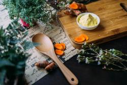 A table with a wooden spoon, cutting board, fresh herbs, and sliced carrots on it. 